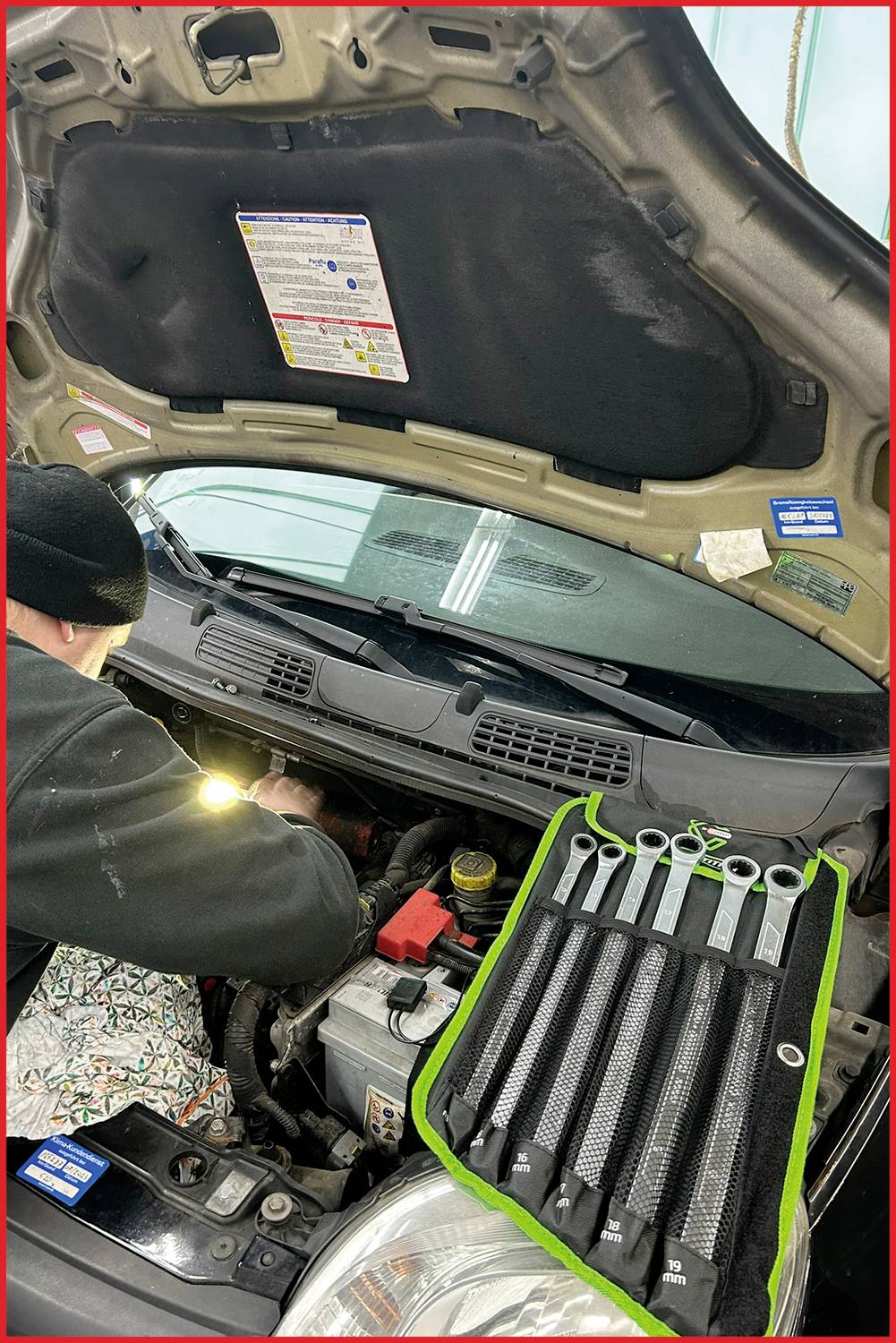 En mekaniker reparerar en bils motor med motorhuven upplyft. En uppsättning skiftnycklar ligger på motortäcket.