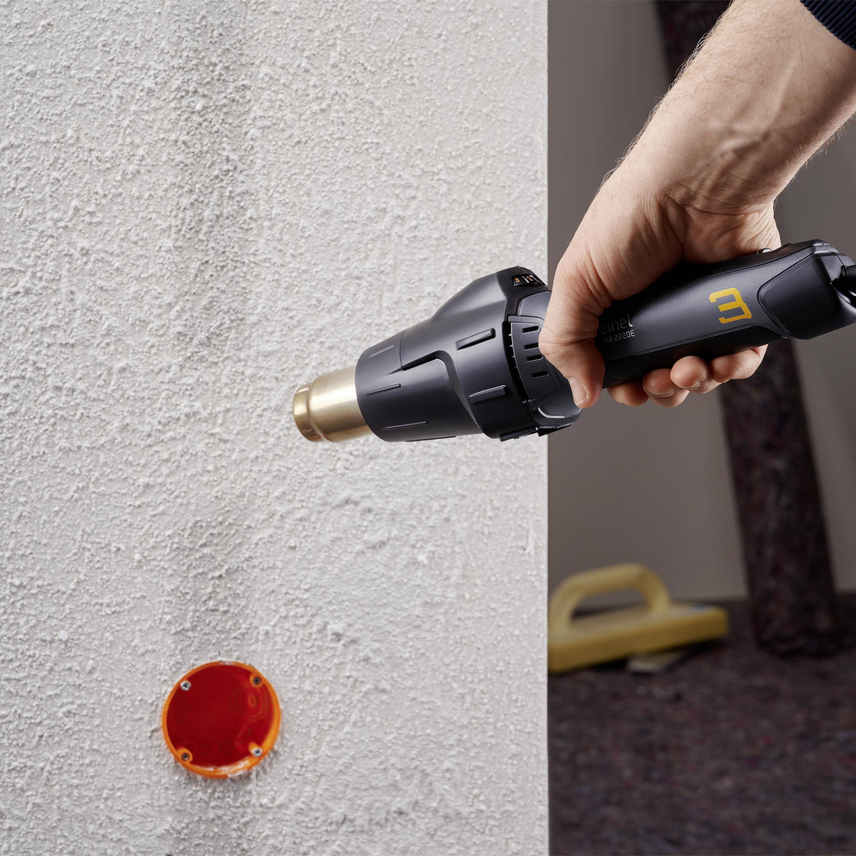 En hand håller en varmluftspistol och värmer ett hål i en grå vägg, sannolikt för att förbereda en installation.