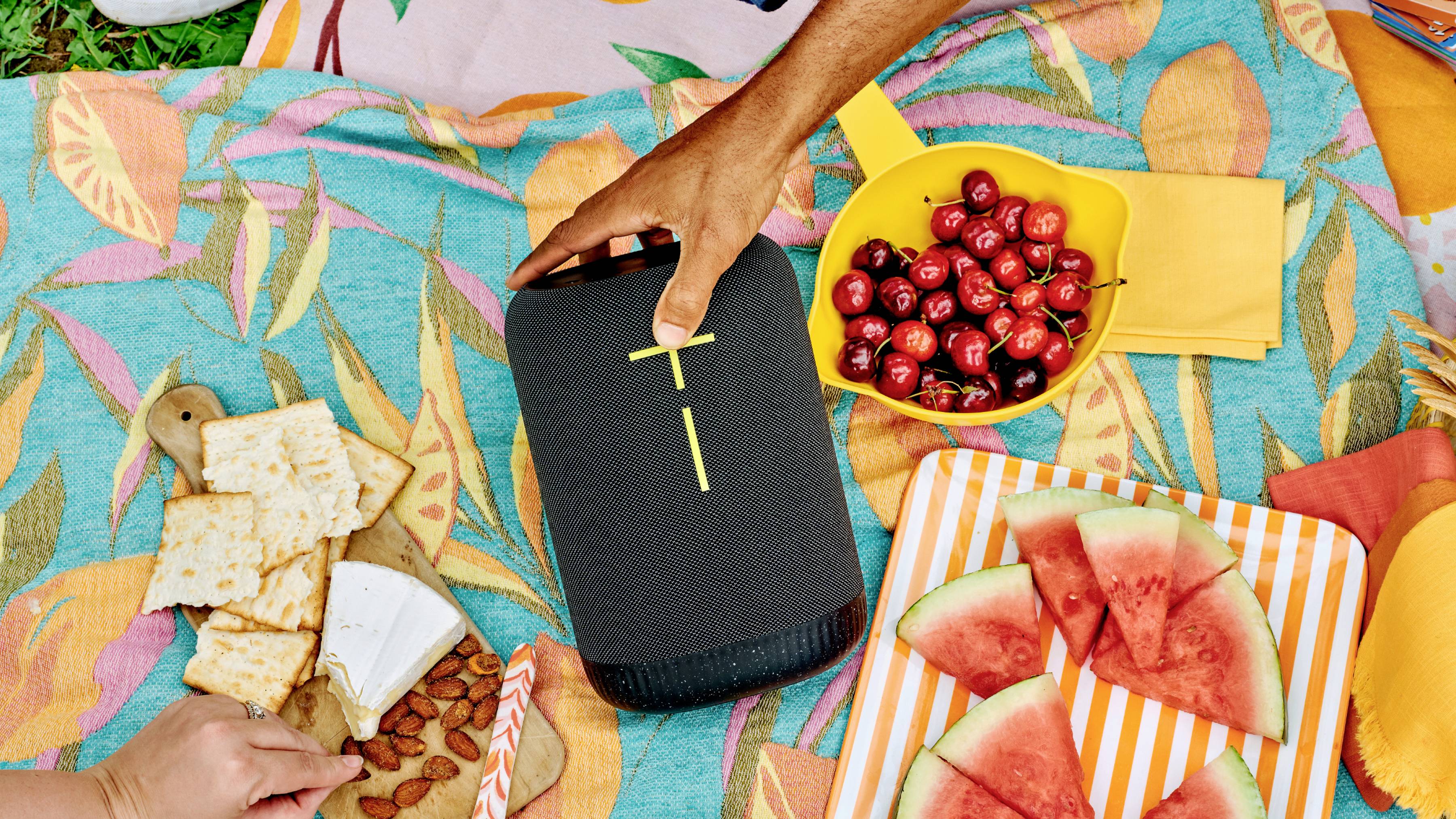 En picknick med melonbitar, körsbär, kex och ost på färgglada filtar utomhus. En hand sträcker sig efter en bärbar högtalare.