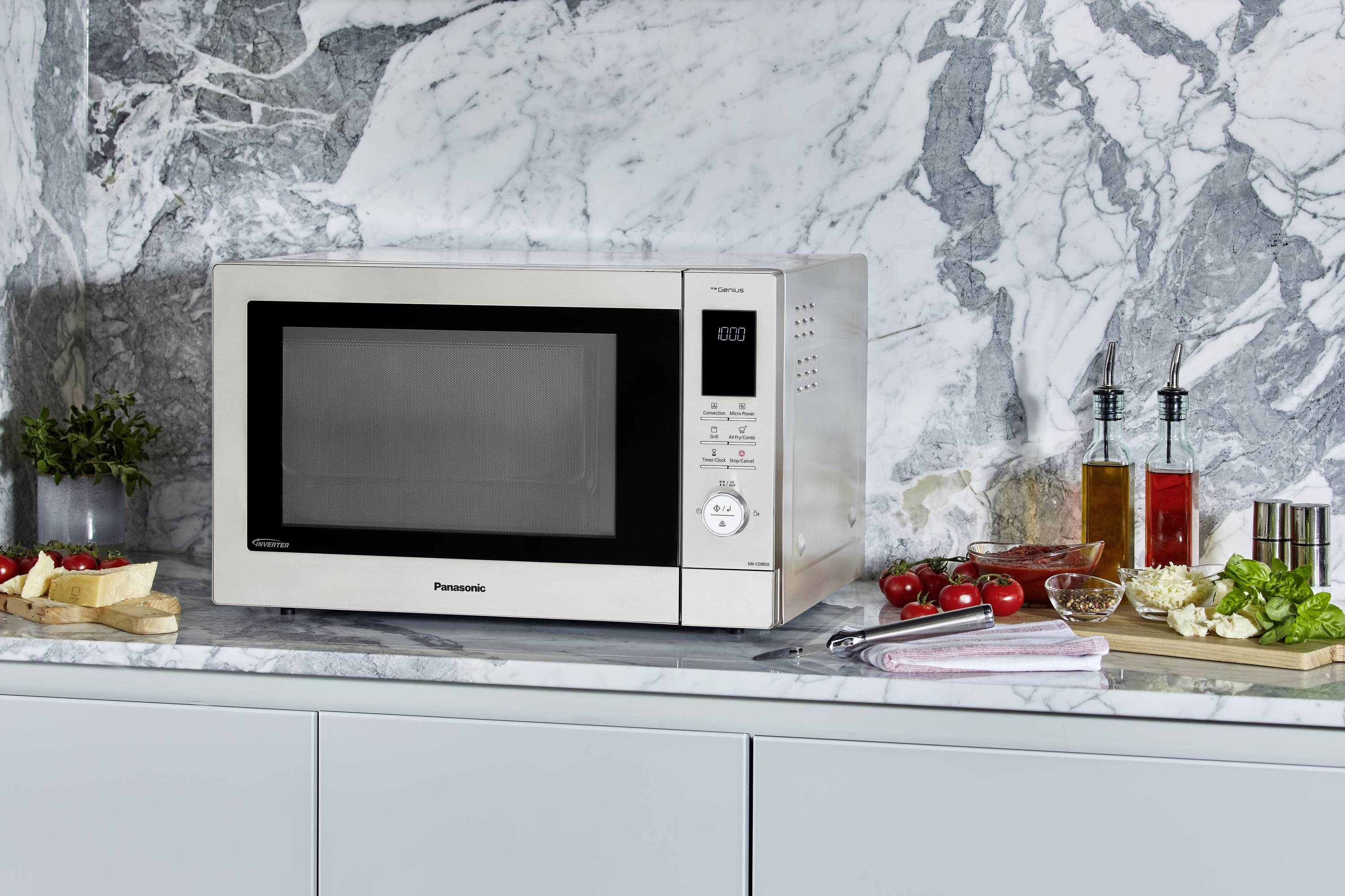 En mikrovågsugn står på en marmorarbetsskiva, omgiven av ingredienser som tomater, ost, broccoli och oljebehållare.