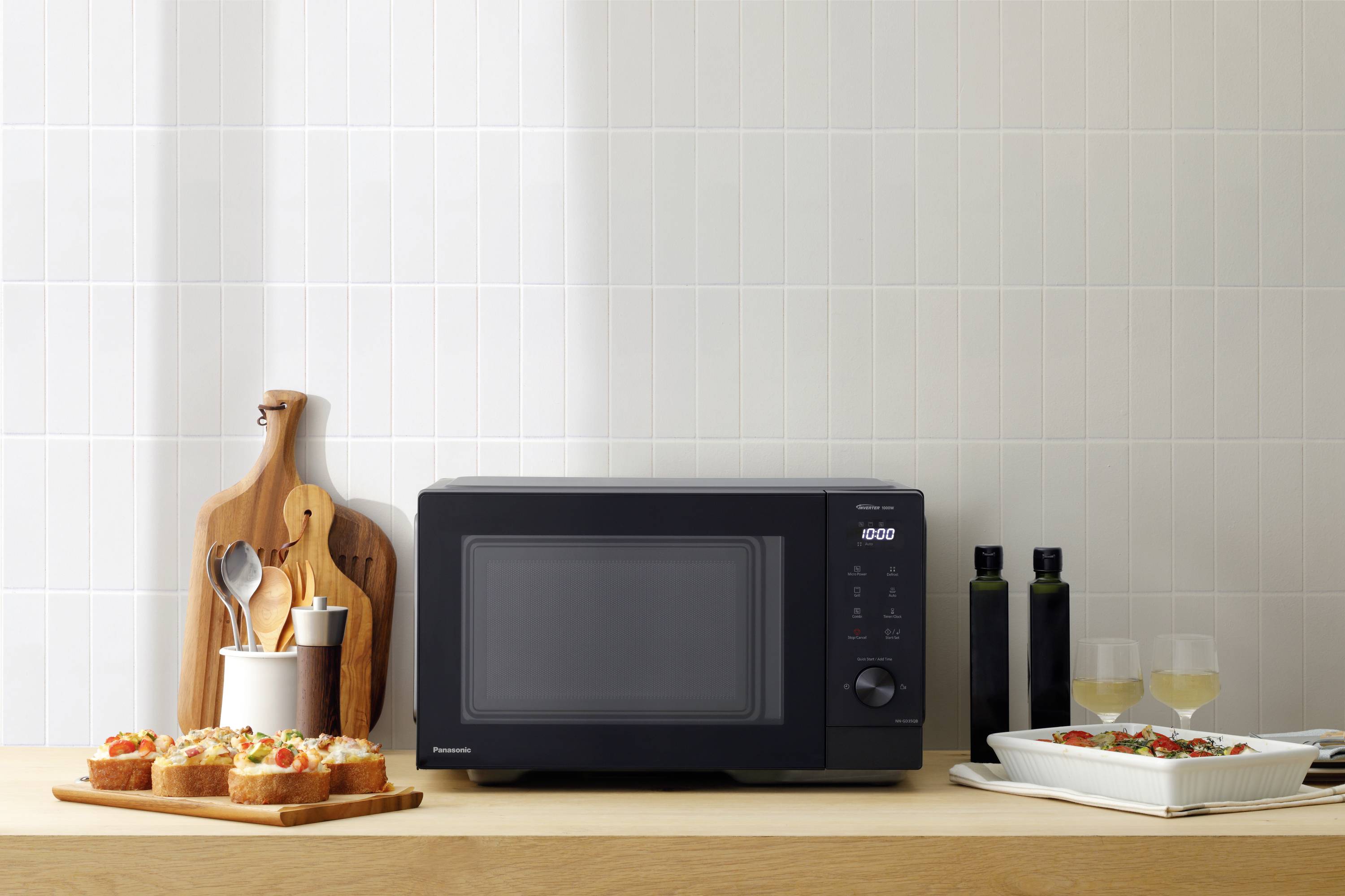 En svart mikrovågsugn står på en träskiva bredvid smörgåsar, två glas med vit vätska och en vit form.