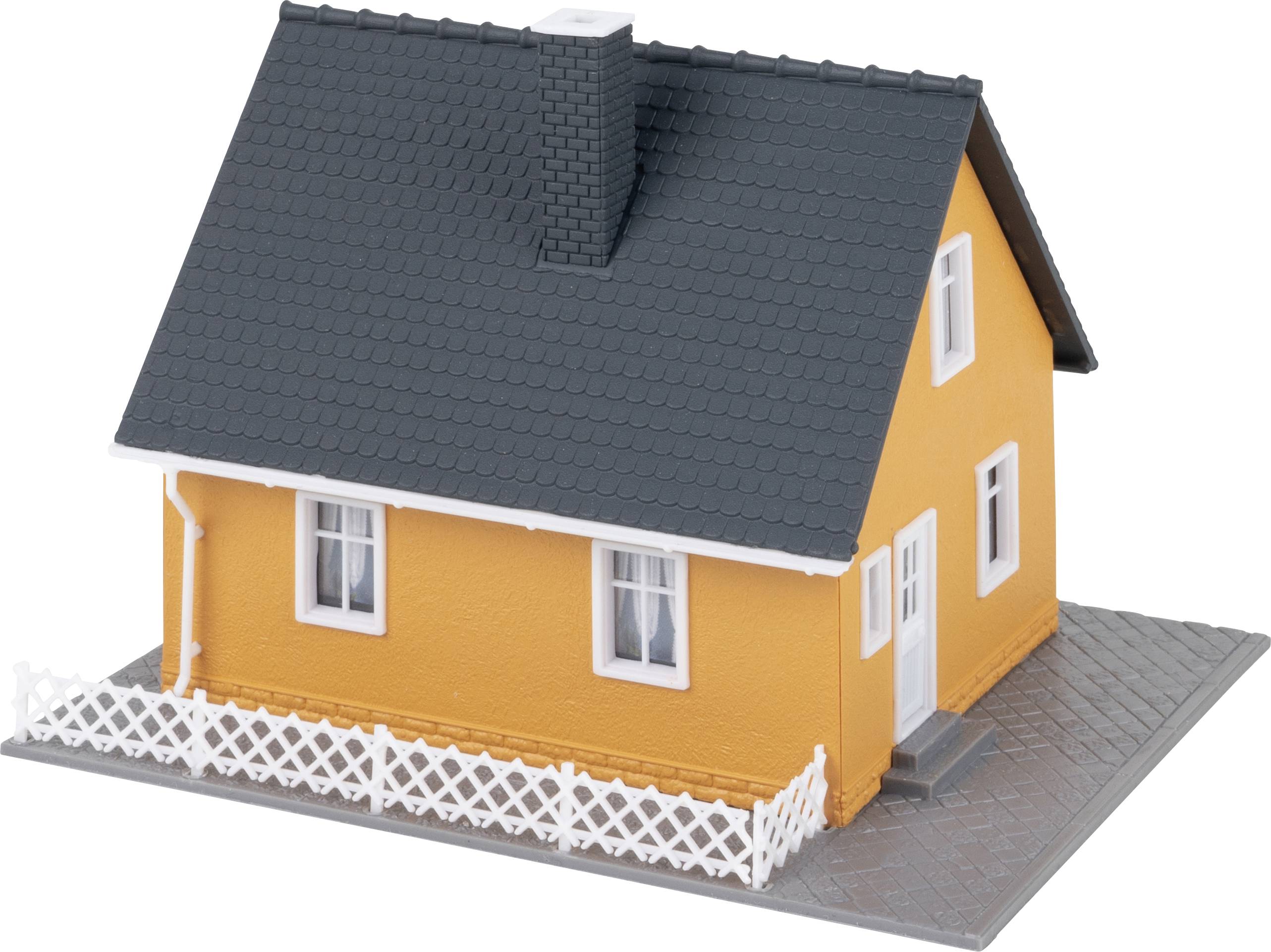 Ett litet modellhus med orange fasad, svarta takpannor och vita fönster, omgivet av ett vitt spjälstaket.