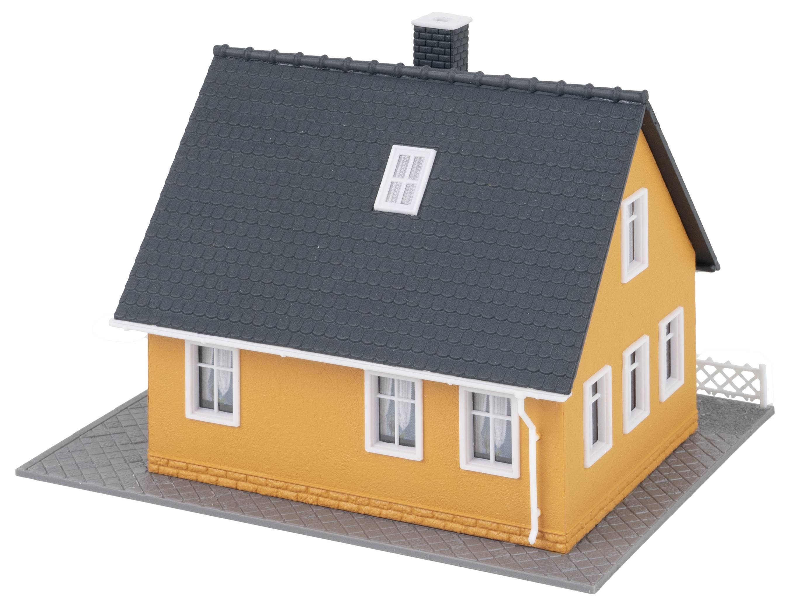 Ett litet modellhus med orangefärgade väggar, grått tak och vita fönsterkarmar. Det står på en grå markyta.