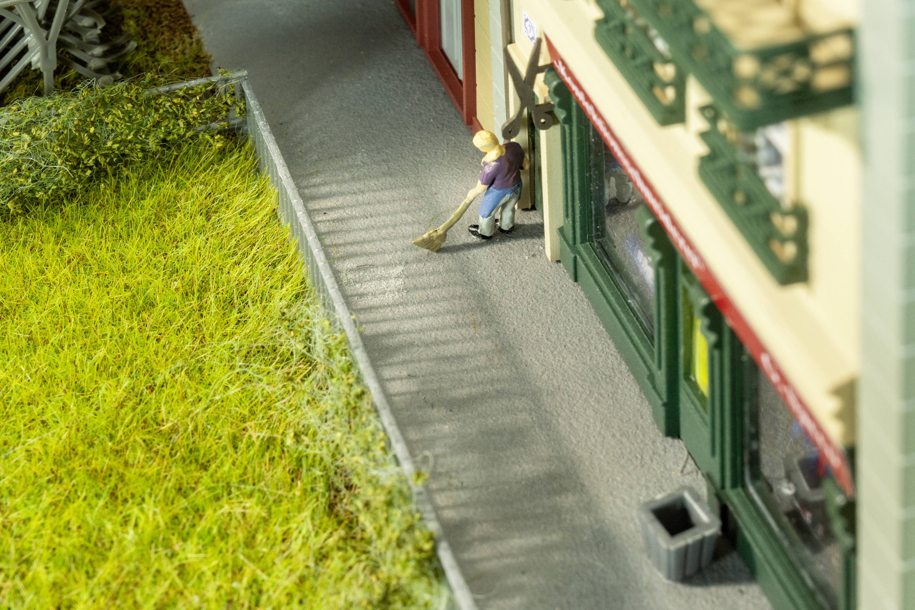 Liten modellfigur sopar trottoaren bredvid en modellbyggnad. En grönyta och en gata ramar in scenen.