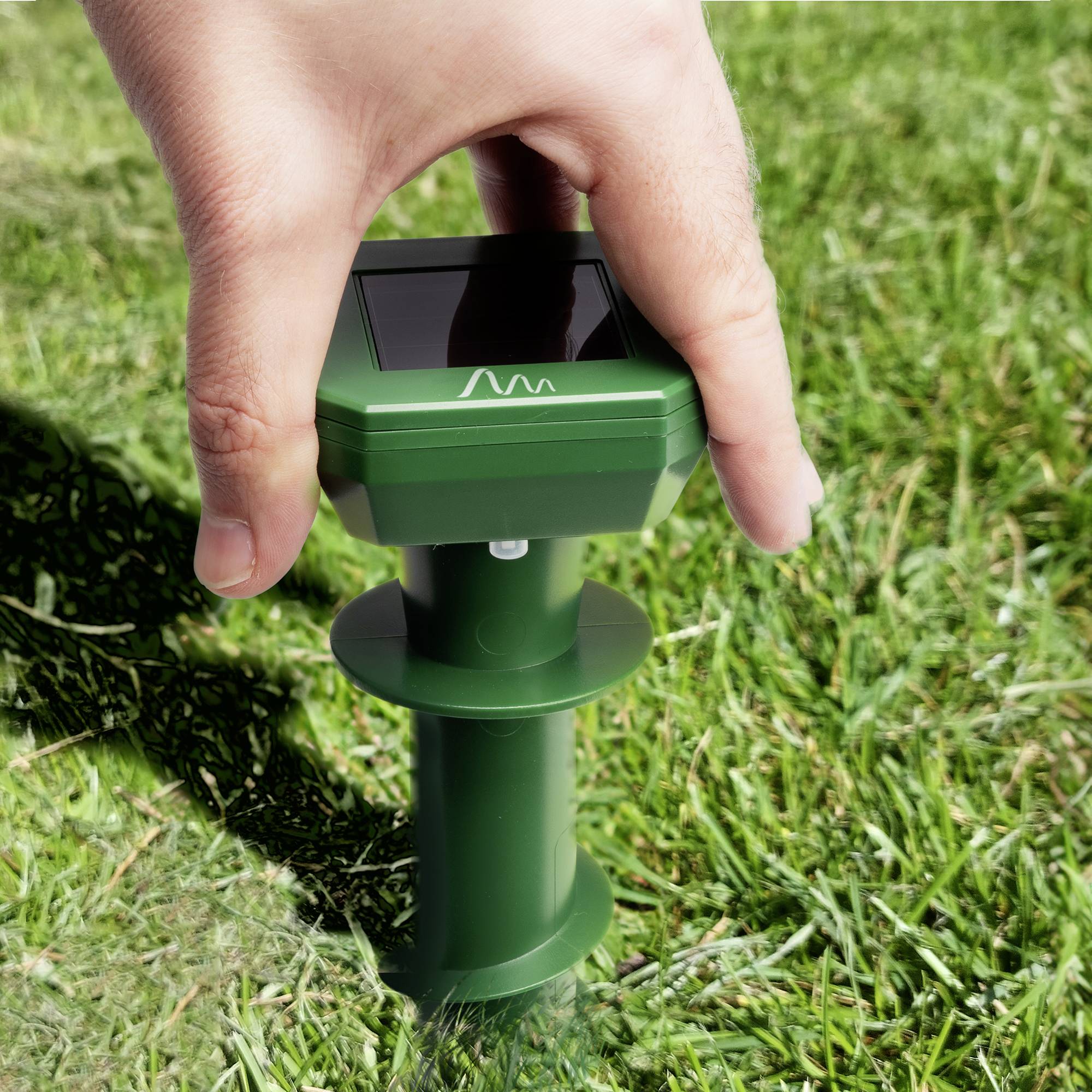 En hand håller en grön, solcellsdriven skrämselapparat för små djur, placerad på en äng.