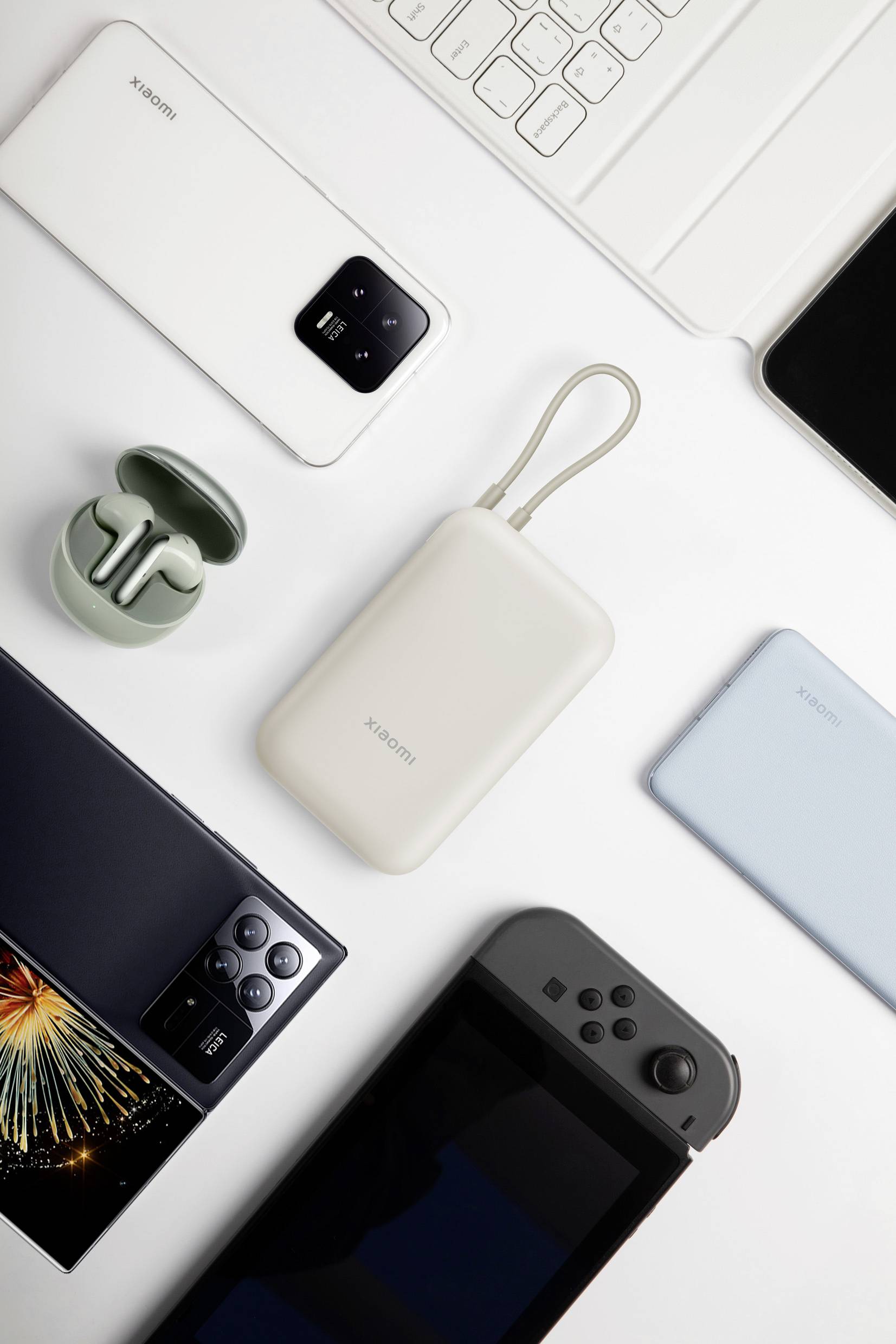 Olika elektroniska prylar inklusive en smartphone, trådlösa hörlurar, powerbank och spelkonsol är arrangerade på en vit yta.