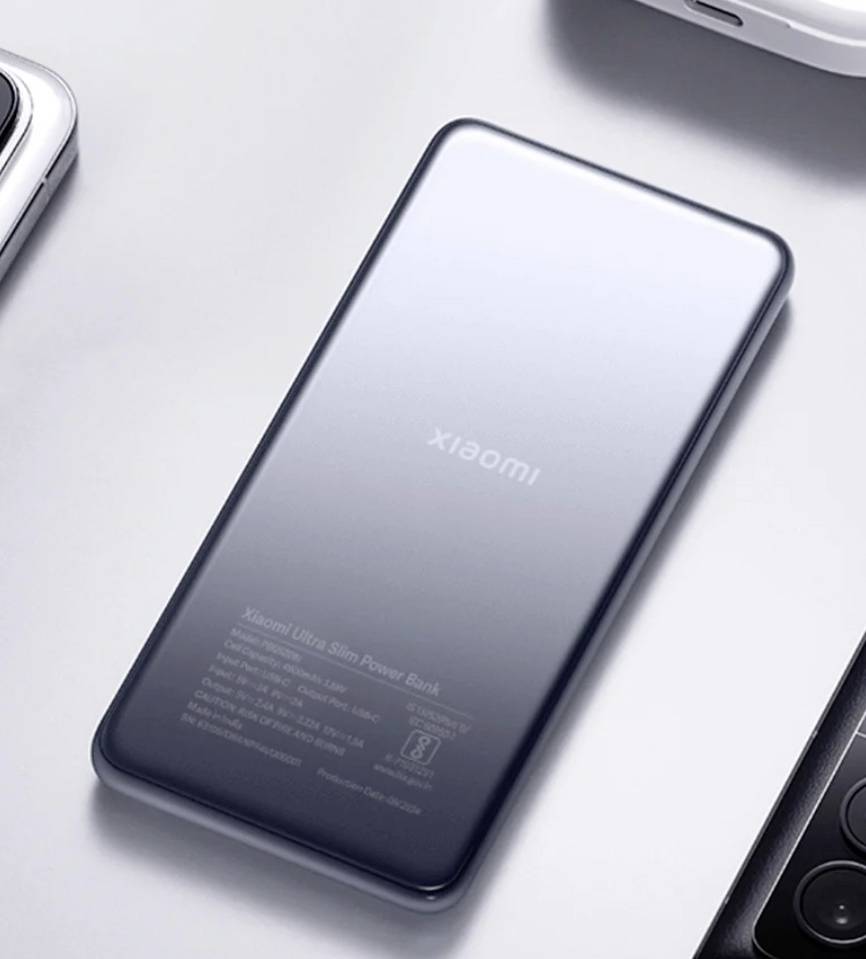 En smal, metallisk powerbank märkt 'Xiaomi' ligger på en ljus yta, omgiven av delar från andra elektroniska enheter.