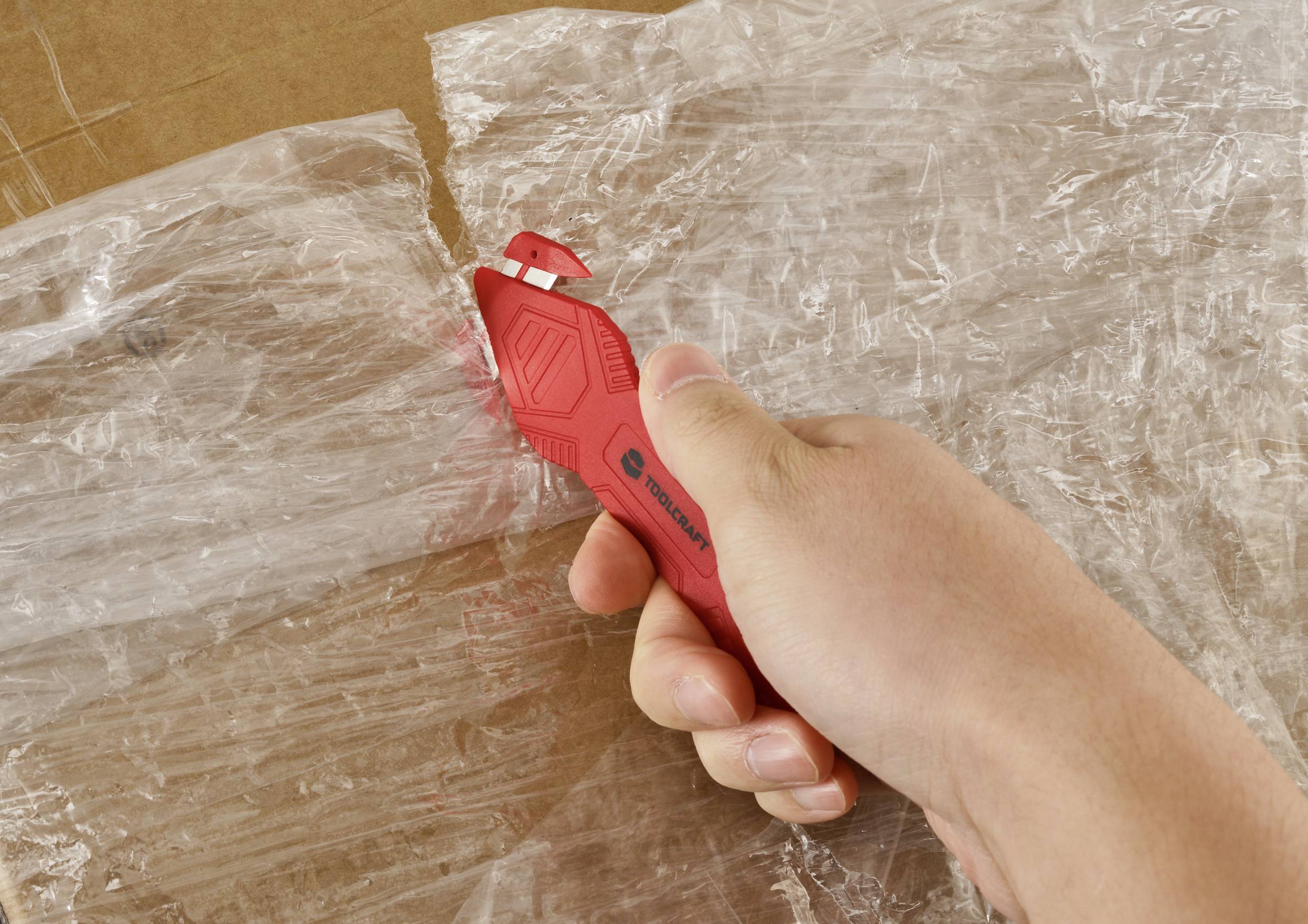 En hand använder en röd universalkniv för att skära igenom genomskinlig plastfolie på en kartong.