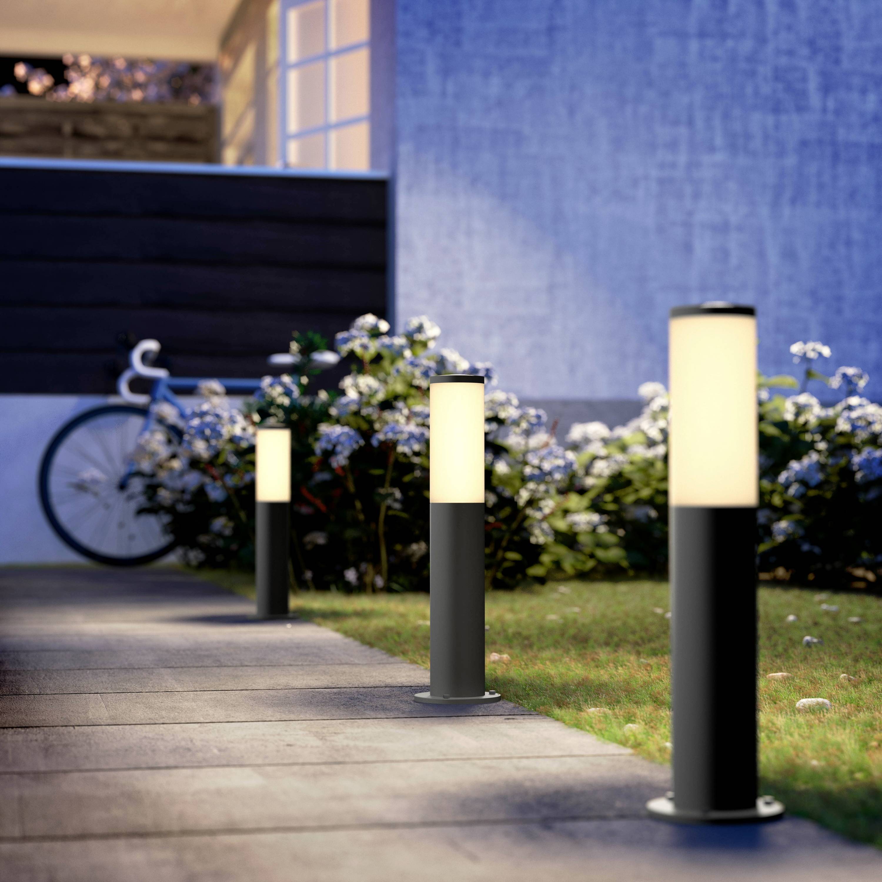 Trädgård med upplysta vägbelysning bredvid en blomrabatt, i bakgrunden en modern byggnad och en cykel.
