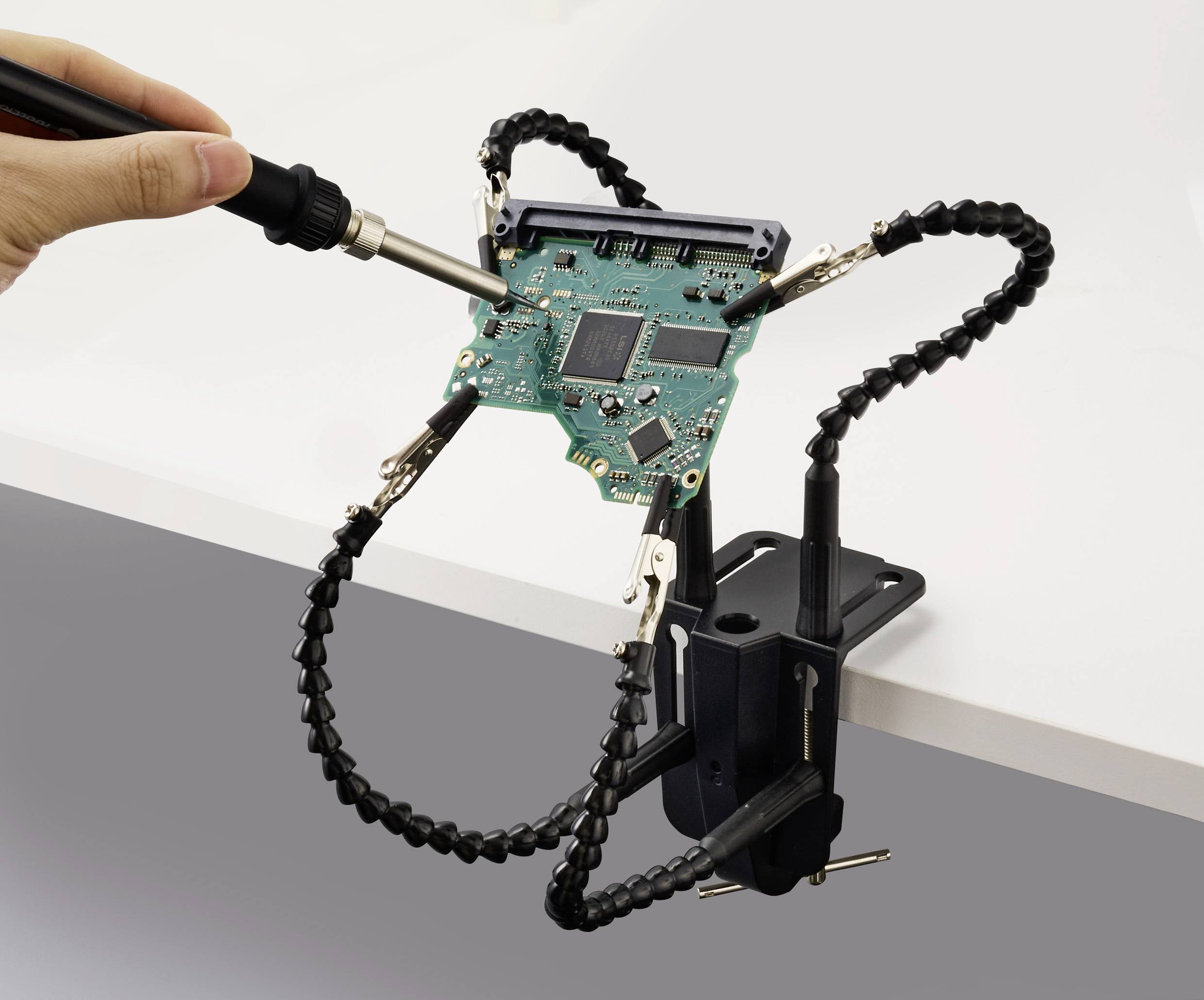 En persons hand löder ett datormoderkort som är fastspänt i en flexibel armklämma, vilket demonstrerar elektronikreparation eller montering.