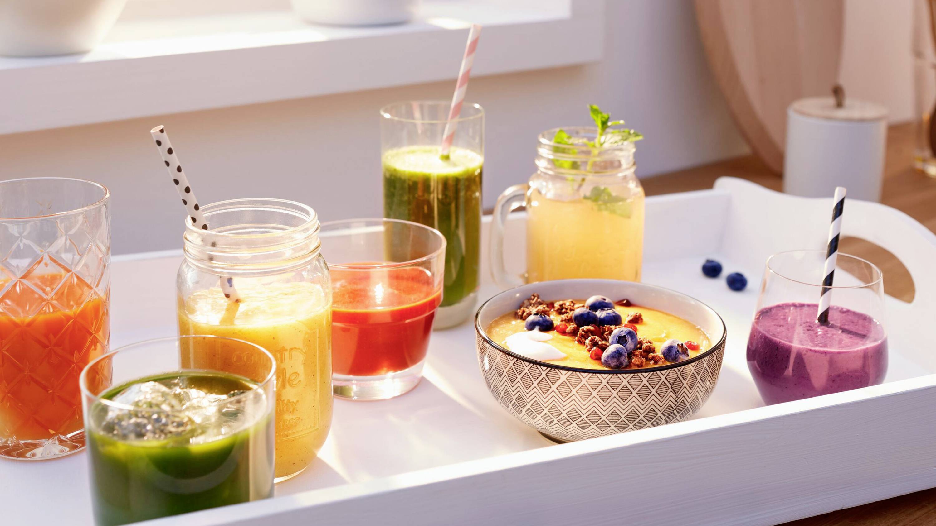 En bricka med färgglada smoothies i olika glas och burkar, och en skål med smoothie toppad med bär, placerad på ett soligt bord.