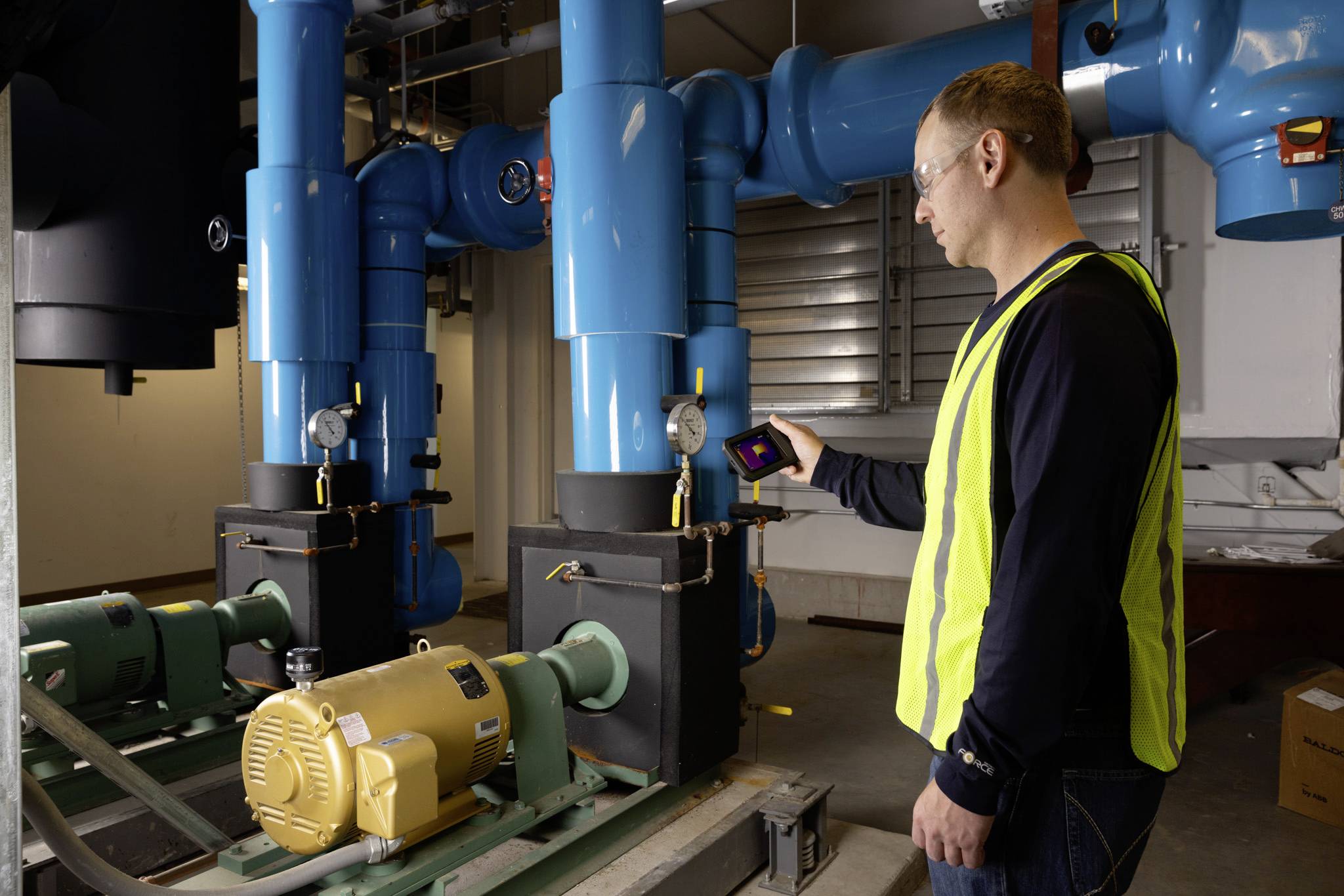 En person i varselväst använder en enhet för att kontrollera blå industriella rör i ett maskinrum, med fokus på mätare och utrustning.
