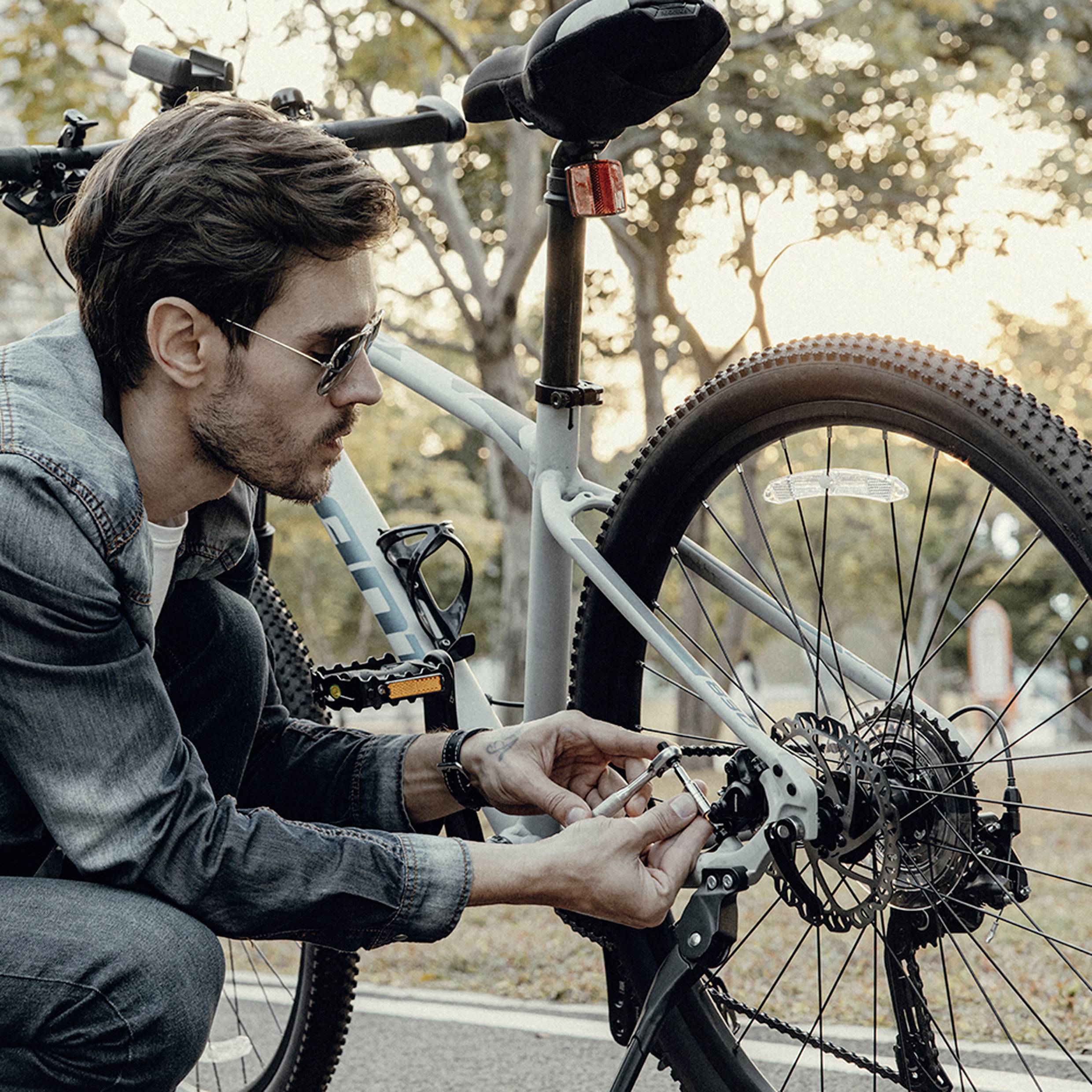 En person i en jeansjacka justerar växlarna på en cykel parkerad i en scenisk parkmiljö, omgiven av träd med höstlöv.
