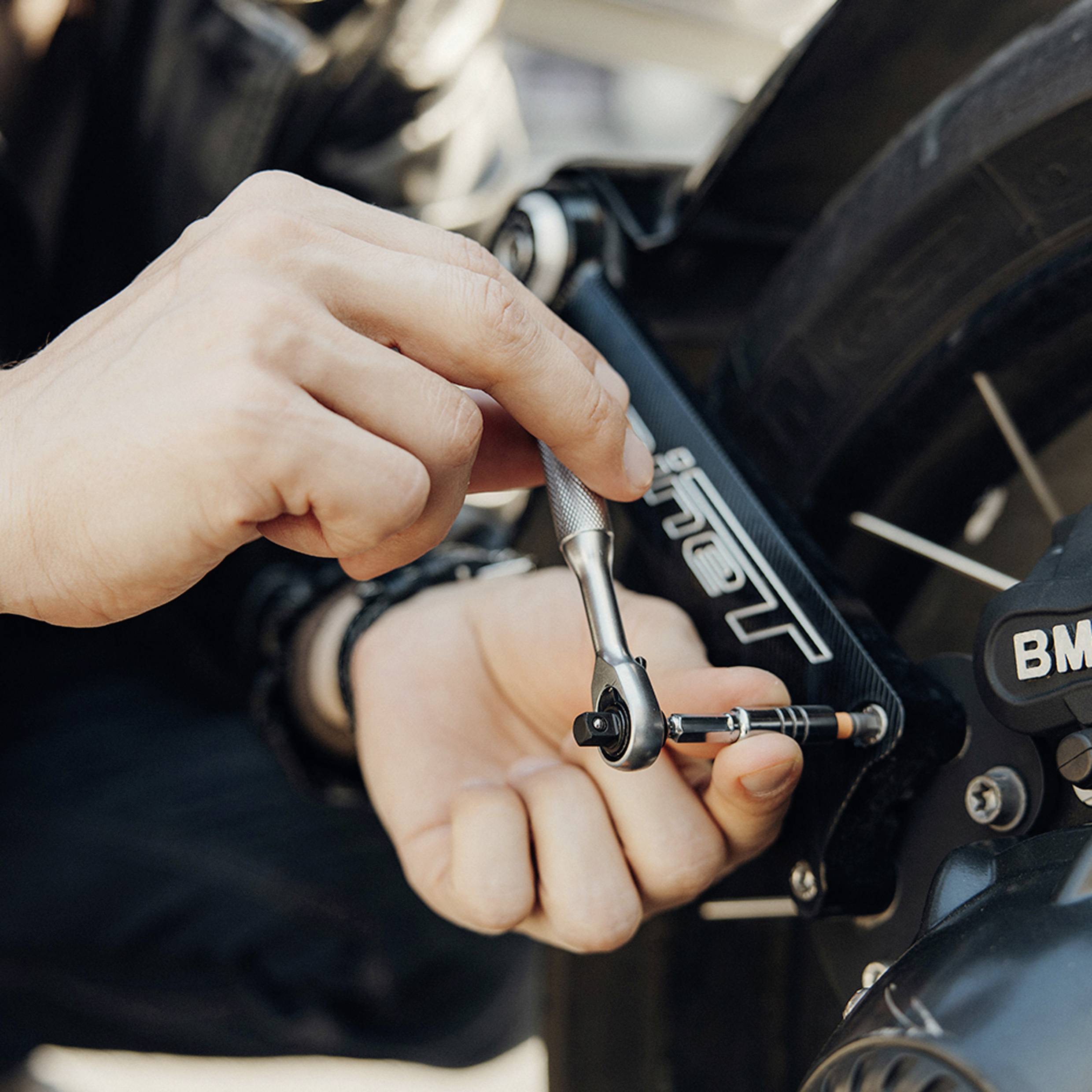 En person använder en skiftnyckel för att justera en motorcykels bakre fotpinnar på en solig dag och fokuserar på precis mekaniskt arbete.