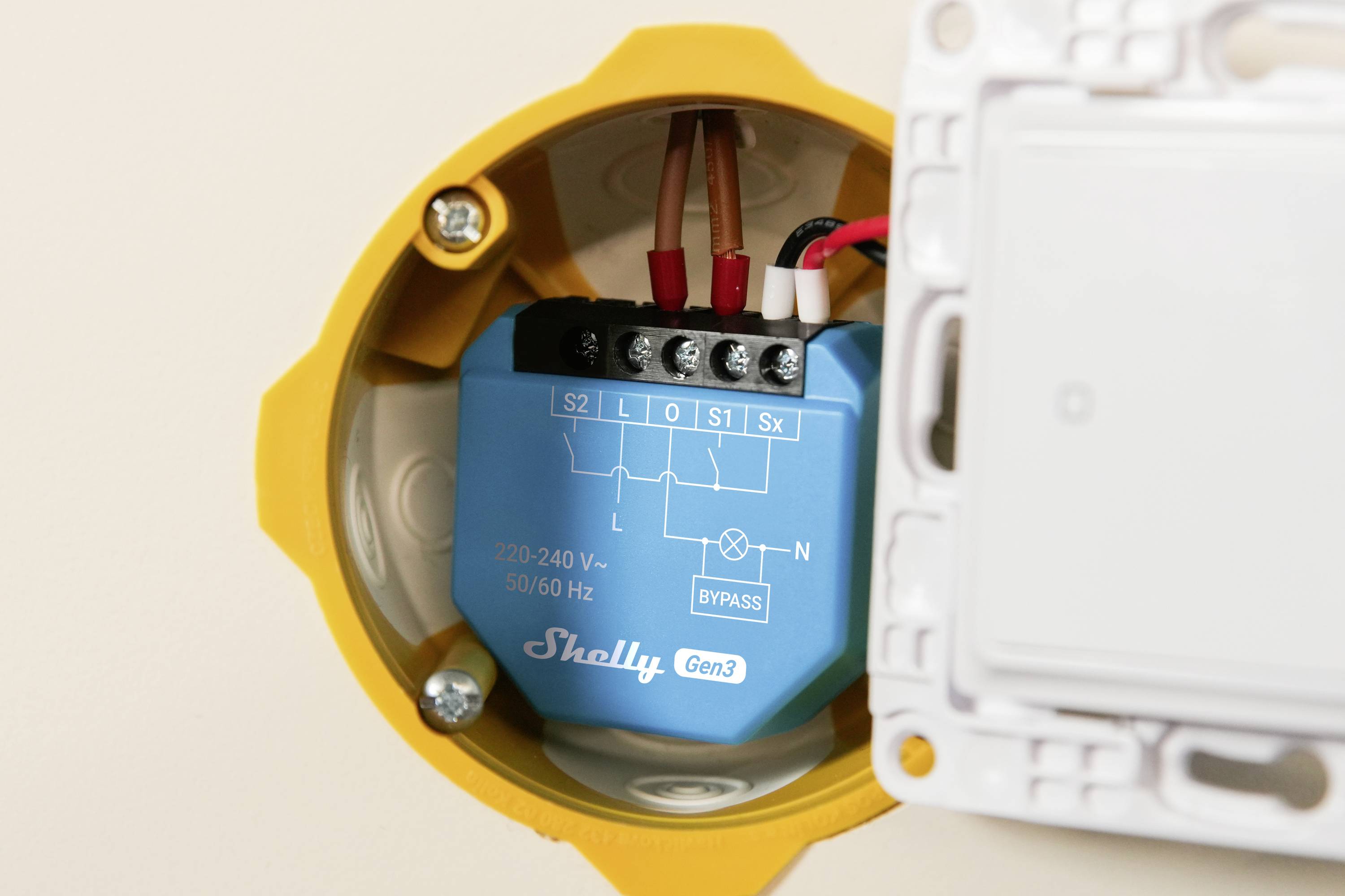 En blå smart enhet märkt 'Shelly Gen3' är installerad inuti ett gult eluttag på väggen. Fyra trådar är anslutna till den.