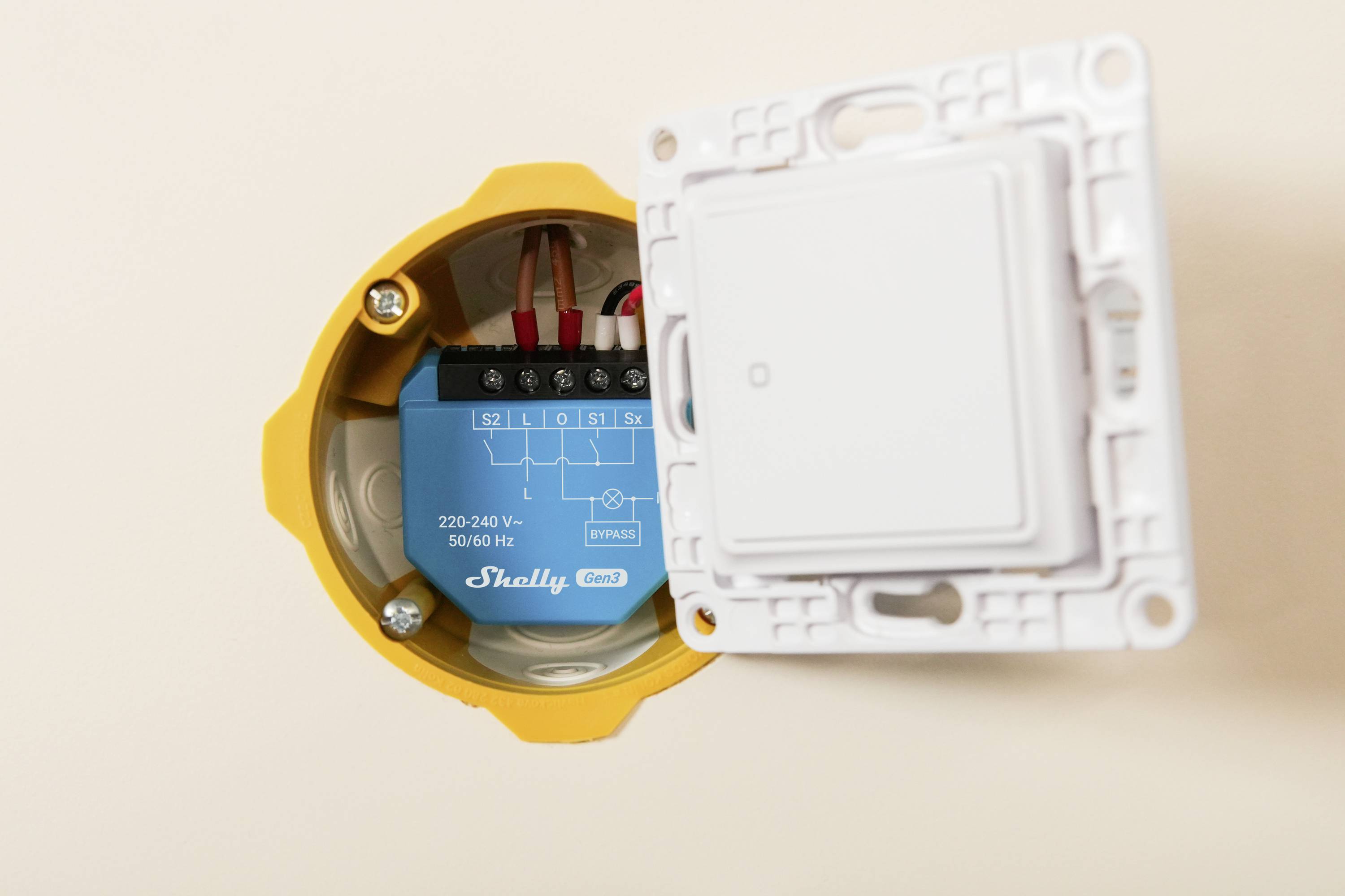En närbild av en elektrisk smart switch installerad i ett vägguttag, som visar kablar och en modul märkt med spännings- och varumärkesinformation.