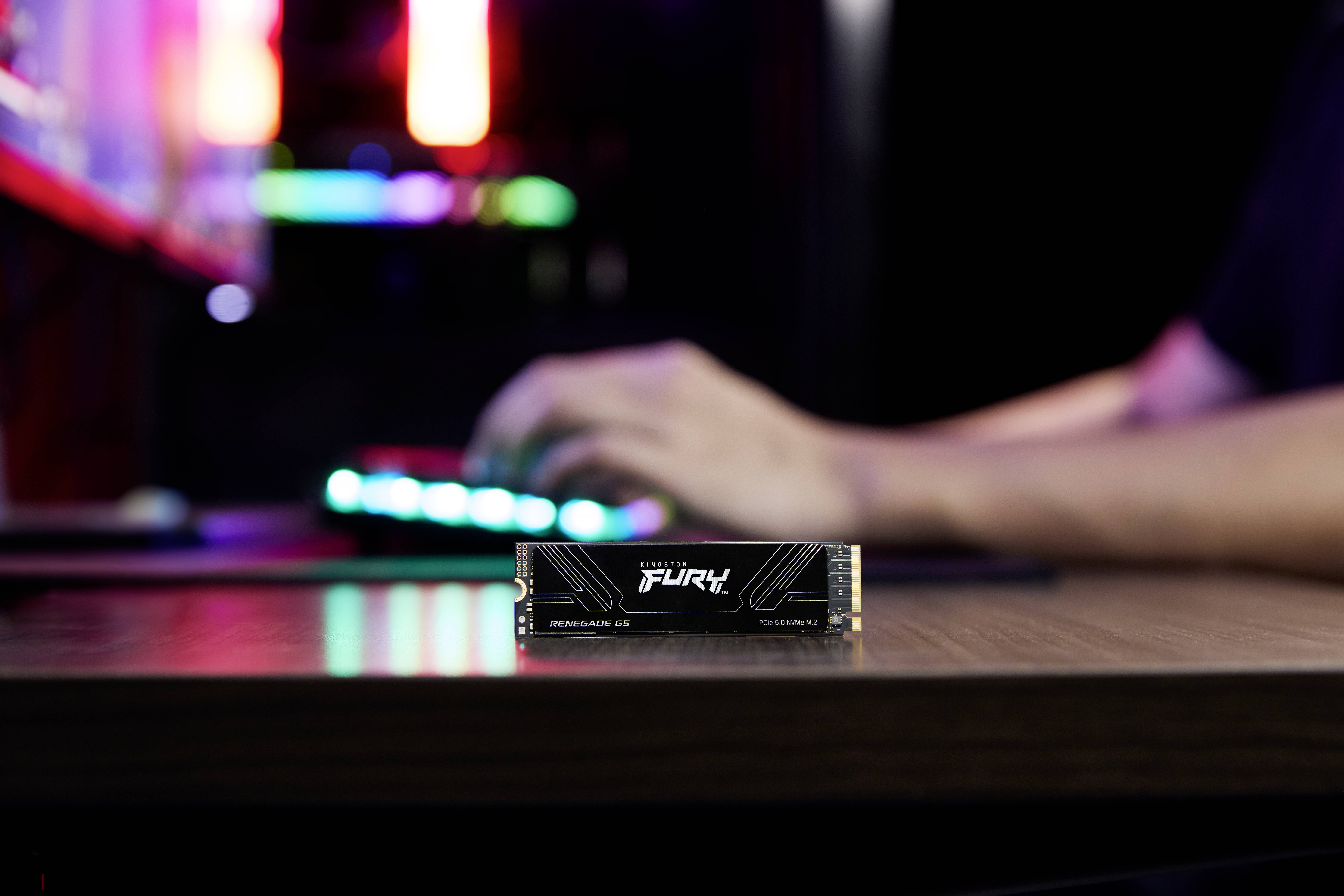 En datentusiast använder ett bakgrundsbelyst tangentbord. I förgrunden är en Kingston Fury Renegade SSD placerad på skrivbordet.