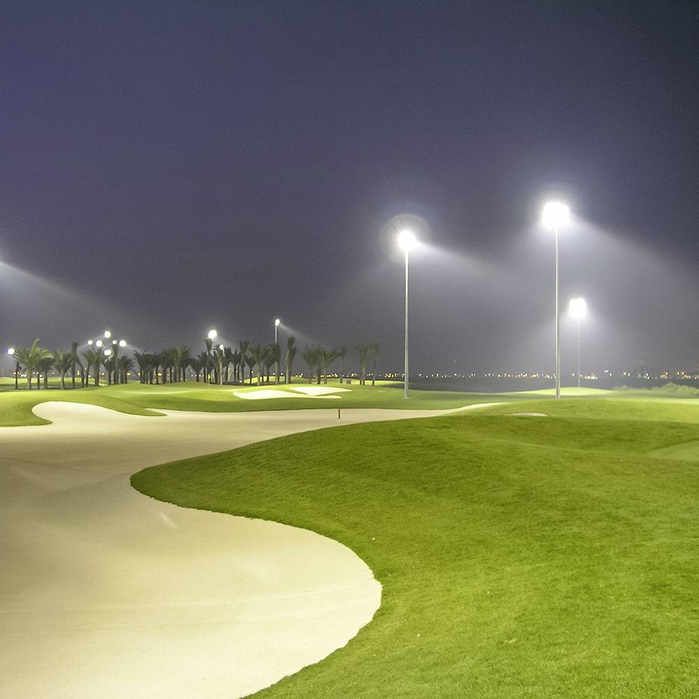 En golfbana upplyst av starka strålkastare nattetid, med frodiga gröna fairways och sandbunkers, omgiven av palmträd.