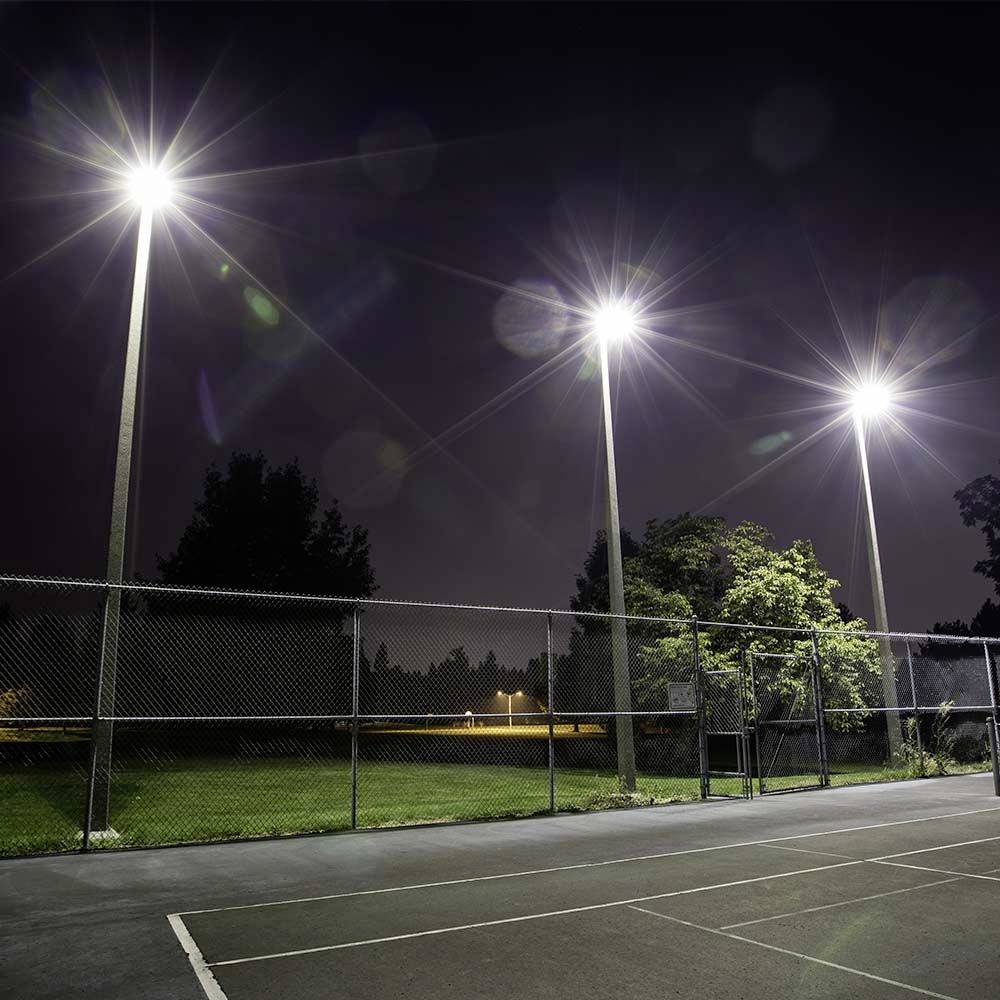 En inhägnad tennisbana upplyst av höga strålkastare nattetid, med omgivande träd delvis synliga i mörkret.