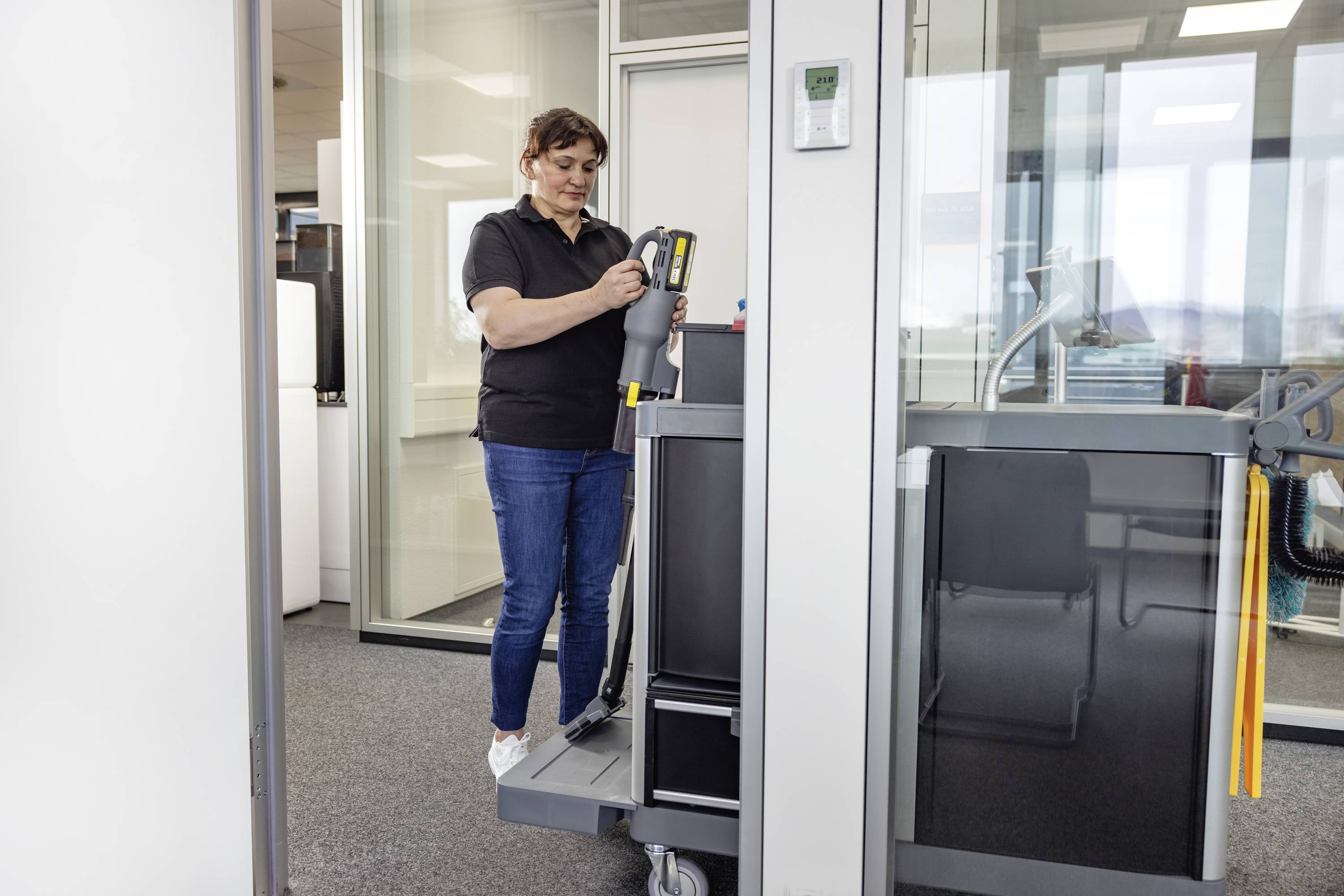 En person i avslappnad klädsel, som står bredvid en städvagn på ett kontor och håller i ett städverktyg, redo att utföra arbetsuppgifter.