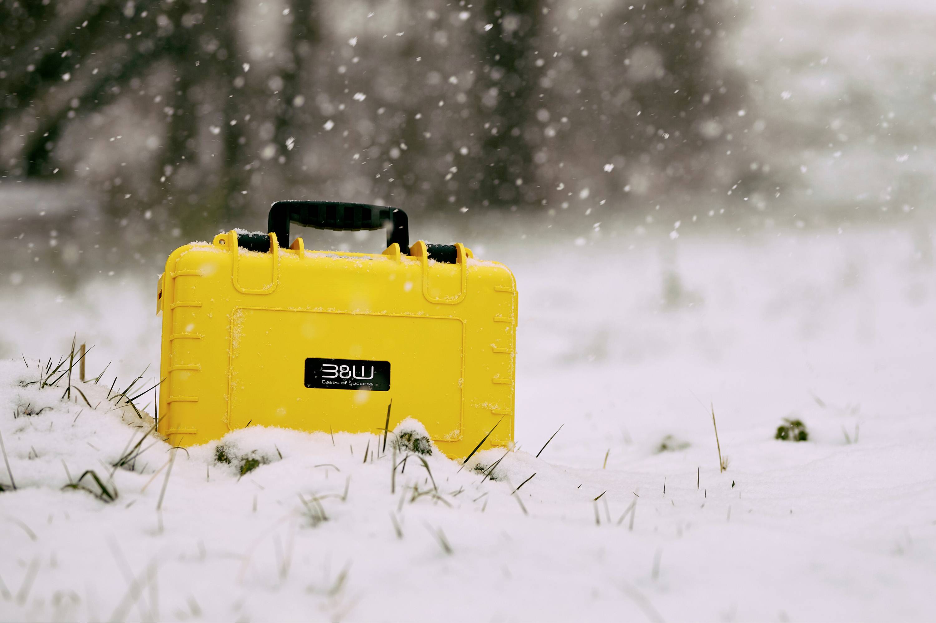 Ett ljusgult fodral med ett handtag sitter på snötäckt mark, omgivet av fallande snöflingor, vilket antyder frilufts- eller utrustningsutrustning.