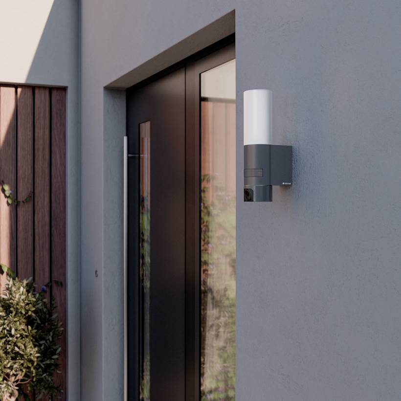 En modern utomhussäkerhetslampa monterad bredvid en dörr på en grå vägg, med ett trästaket och buskage synligt i bakgrunden.