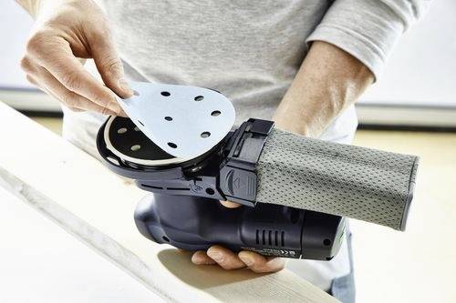 En person i en långärmad tröja fäster en ny slipskiva på en elektrisk slipmaskin. Slipmaskinen har en dammuppsamlingspåse påkopplad.