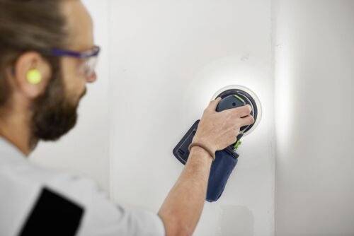 En person sliper en vit vägg med en rund elektrisk slipmaskin, bär hörselskydd och ögonskydd, med fokus på att jämna ut ytan.