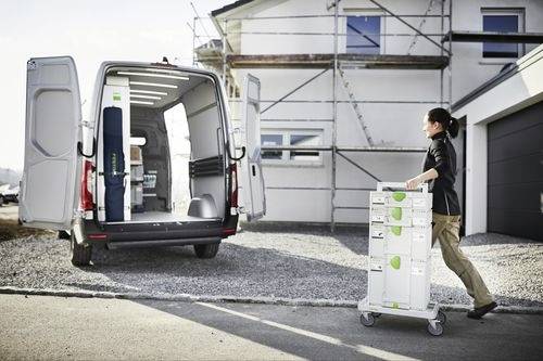En person skjuter en vagn fylld med lådor mot en öppen skåpbil parkerad intill ett hus under byggnation, vilket tyder på en leverans eller flytt.