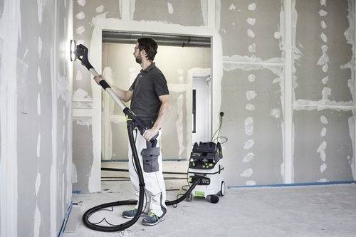 En person slipar en vägg med en elektrisk slipmaskin med långt handtag, ansluten till en dammsugare, i ett delvis färdigställt rum med gipsskivor.