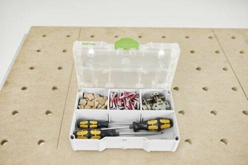 En öppen plastbehållare med fack som innehåller träbitar, röda kopplingar, metallclips och två skruvmejslar på en perforerad skiva.