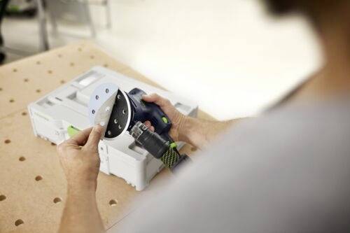 En person justerar sandpapper på en elektrisk slipmaskin och förbereder sig för en träbearbetningsuppgift på en perforerad arbetsbänk i en verkstadsmiljö.
