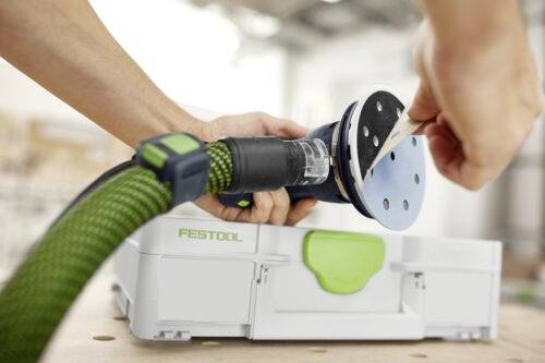 Person som justerar slippapperet på en orbital slipmaskin, med en Festool verktygslåda i bakgrunden, vilket tyder på en snickeri- eller verkstadsmiljö.