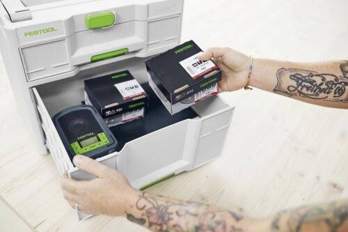 En person laddar en låda med Festool batteriladdare och batterier. Armen har tatueringar, och lådan och produkterna är märkta.