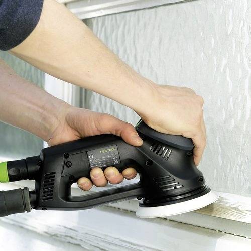 En person använder en elektrisk slipmaskin på en träfönsterram och jämnar ut ytan. Slipmaskinen är svart med en synlig nätsladd.