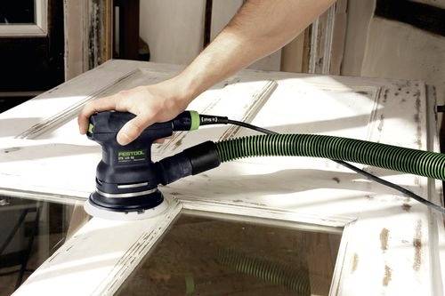 En person använder en elektrisk slipmaskin på en vit träddörr och jämnar ut ytan. Scenen antyder hemmafixande eller träarbete.