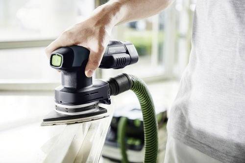 En person som använder ett elslipverktyg med en dammuppsamlingsslang ansluten, i ett ljust rum intill ett fönster.