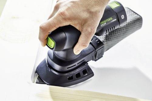 En hand som använder en elektrisk slipmaskin på en träyta och jämnar till kanten. Verktyget är svart och grönt med ergonomiska handtag.