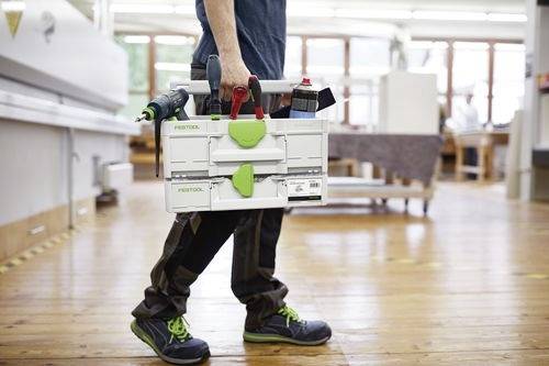 En person bär en Festool-verktygsväska i en verkstad, vilket antyder aktivt arbete eller förberedelse för en uppgift med olika verktyg inuti.