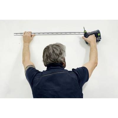 En person mäter en vit vägg med ett måttband och håller i en borrmaskin, förberedd att montera eller installera något.