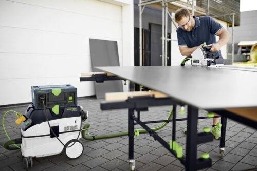 En man använder en cirkelsåg för att såga ett stort skivmaterial på en arbetsbänk utomhus, med ett vakuumsystem anslutet för dammsugning.