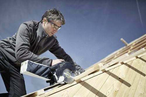 En person som använder en cirkulär såg för att såga träplankor på ett sluttande tak, iklädd skyddsglasögon och utomhuskläder under en klarblå himmel.