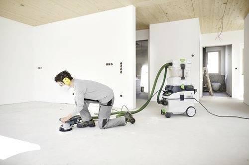 En person som bär skyddsutrustning använder en golvslipmaskin ansluten till en industriell dammsugare i ett vitt, möblerslöst rum.
