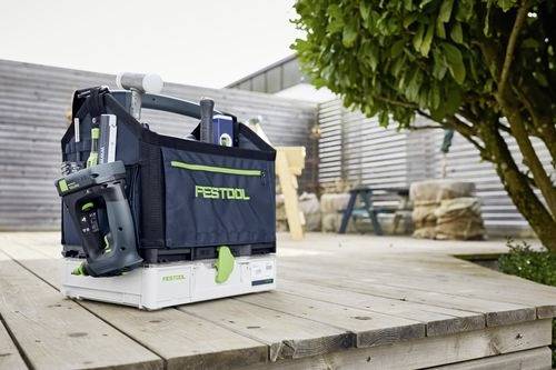 En låda med Festool-verktyg står på ett trätrall med ett träd till höger. En modern byggnad och stensocklar syns i bakgrunden.