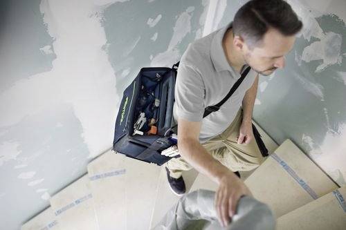En person som bär en verktygstaske stiger upp för en delvis målad trappa, vilket tyder på pågående renoverings- eller underhållsarbete i ett hem.