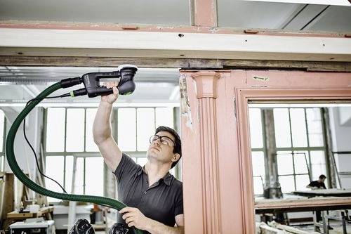 En man använder en elektrisk slipmaskin för att renovera en stor trädörrkarm i en verkstad.