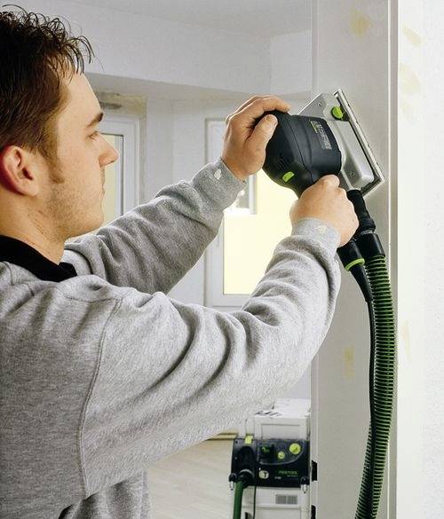 En person som använder en elektrisk slipmaskin för att jämna ut en värnghörn; dammsugare ansluten för att minska dammspridningen.