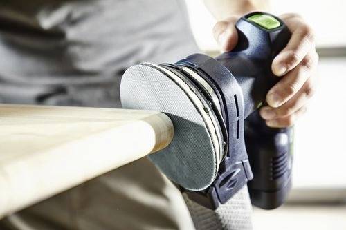En person som använder en elektrisk slipmaskin för att jämna till kanten på en träyta, med fokus på hantverksskicklighet och precision.