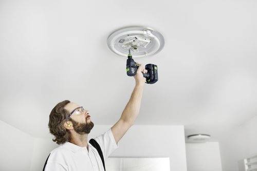En person som installerar en taklampa med hjälp av en sladdlös borrmaskin, bär hörselskydd och skyddsglasögon i ett ljust rum.