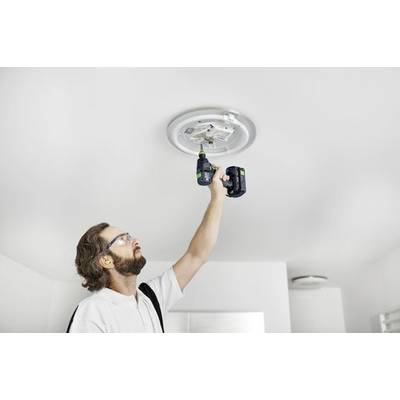 En person som installerar en taklampa med hjälp av en sladdlös borrmaskin, bär hörselskydd och skyddsglasögon i ett ljust rum.